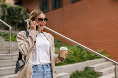 Smiling woman office worker is talking by phone while standing on modern building background 