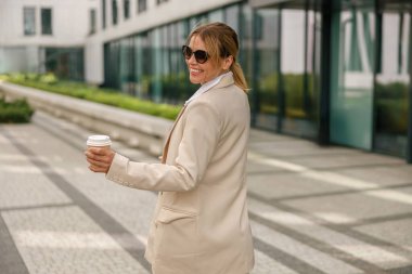 Kahveli gülümseyen kadın ofis çalışanı, teneffüste modern bina arka planında yürüyor.