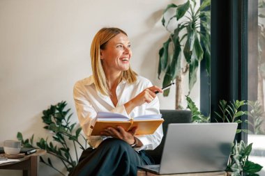 Gülümseyen kadın girişimci dizüstü bilgisayarda çalışıyor, samimi iç mekan ile ilgili notlar alıyor.