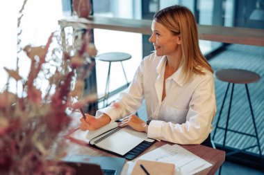 Gülümseyen kadın girişimci dizüstü bilgisayarda çalışıyor, samimi iç mekan ile ilgili notlar alıyor.