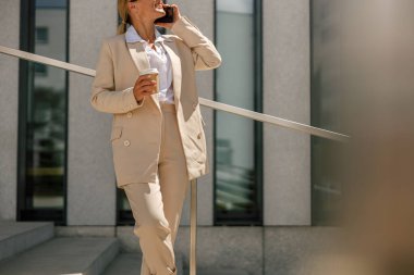 Gülümseyen kadın ofis çalışanı modern binanın arka planında duruyor ve kahve içiyor.