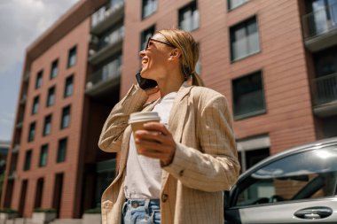 Gülümseyen kadın ofis çalışanı modern binanın arka planında duruyor ve kahve içiyor.