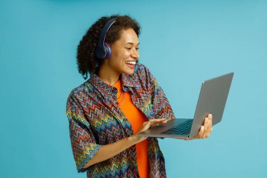 Gülümseyen kadın kulaklıklı, dizüstü bilgisayarda çalışırken mavi stüdyo arka planında duruyor.