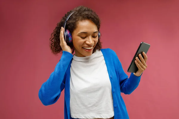 stock image Cheerful woman in earphones holding mobile phone and listen music standing on red studio background 