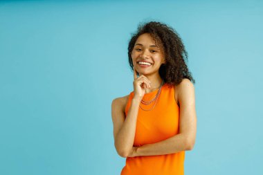 Turuncu elbiseli gülümseyen kadın yanağına dokunuyor ve mavi arka plandaki kameraya bakıyor.