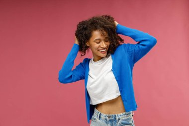 Kırmızı stüdyo arka planında dikilirken gülümseyen ve saçlarına dokunan pozitif kadın. 