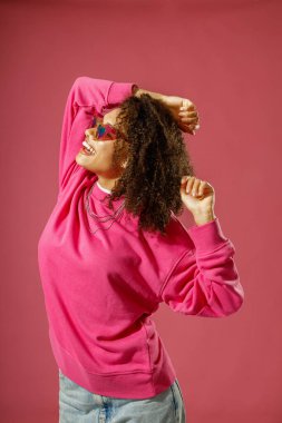 Pembe stüdyo arka planında duran, süveterli ve kalp şeklinde gözlüklü gülümseyen kadın.