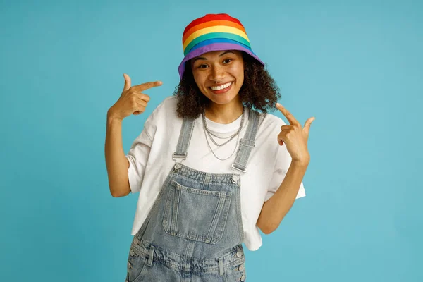 青いスタジオの背景の上に立っている自分自身を指を指す虹の帽子で誇りに思える笑顔の女性 — ストック写真