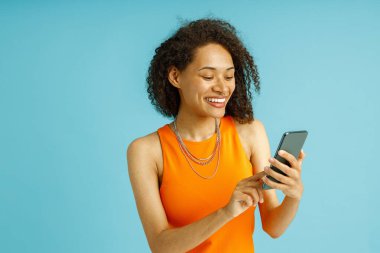 Turuncu elbiseli güzel kadın mavi stüdyo arka planında dururken cep telefonu kullanıyor.