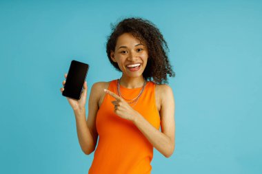 Gözlüklü güzel kadın cep telefonunu işaret ediyor. Mavi stüdyo arka planında duruyor.