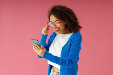 Pembe stüdyo arka planında dikilirken cep telefonuna bakan pozitif kadın