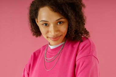 Güzel bir kadın pembe stüdyo arka planında dururken gülümseyerek kameraya bakar.