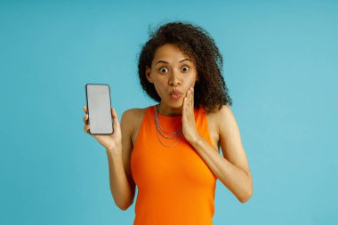 Şok olmuş bir kadın kameraya bakarken yanağına dokunuyor ve telefonu mavi stüdyo arka planında tutuyor.