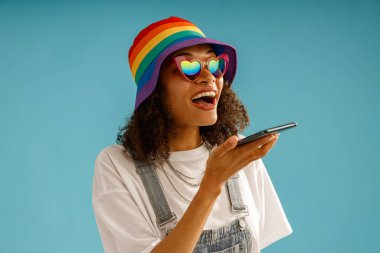 Gökkuşağı şapkalı neşeli kadın mavi stüdyo arka planında telefonla sesli mesaj kaydediyor.