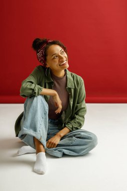 Çekici bir kadın yerde oturuyor ve kırmızı stüdyo arka planına gülümsüyor.