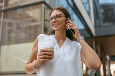 Kahve fincanı olan güzel bir iş kadını modern binanın arka planında duruyor ve telefonla konuşuyor.