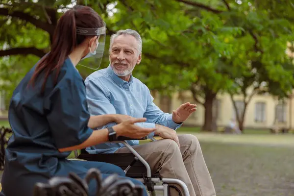 Talkative aged man, recovering patient in wheelchair having conversation with his nurse in protective face shield, resting together in the park near hospital. Support, rehabilitation concept