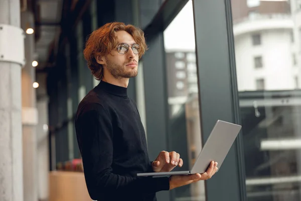 労働日の間にオフィスで立っているラップトップで働く眼鏡のハンサムな男性営業マネージャー ストック写真