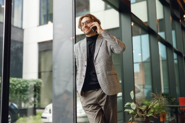 Cep telefonuyla konuşan yakışıklı bir işadamı pencerenin yanında duruyor ve yüzünü çeviriyor.