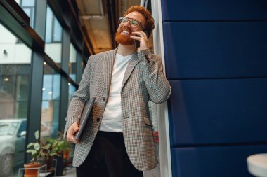 Gözlüklü, sakallı, serbest çalışan bir erkek modern çalışma alanında telefonla konuşuyor.