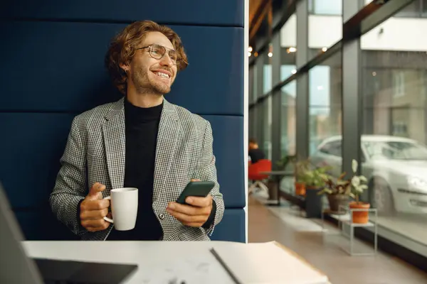 笑顔のビジネスマンは コワーキングの窓の近くに座っている間 携帯電話とコーヒーを飲む — ストック写真
