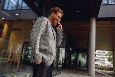 Gözlüklü, sakallı, serbest çalışan bir erkek modern çalışma alanında telefonla konuşuyor.