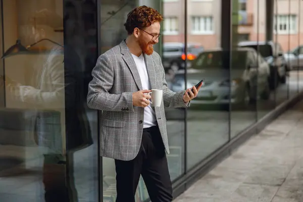 Smiling businessman holding phone while standing near office during break time. High quality photo
