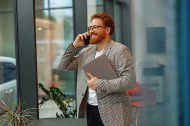 Bilgisayarlı gülümseyen girişimci pencerenin yanındaki modern ofiste telefonla konuşuyor