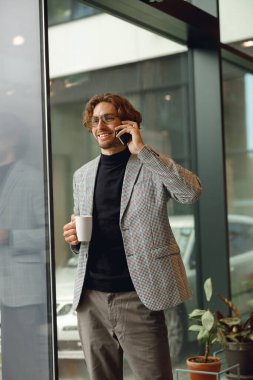 Havalı erkek serbest çalışan cep telefonuyla konuşuyor. Pencerenin yanında duruyor ve kahve içiyor.