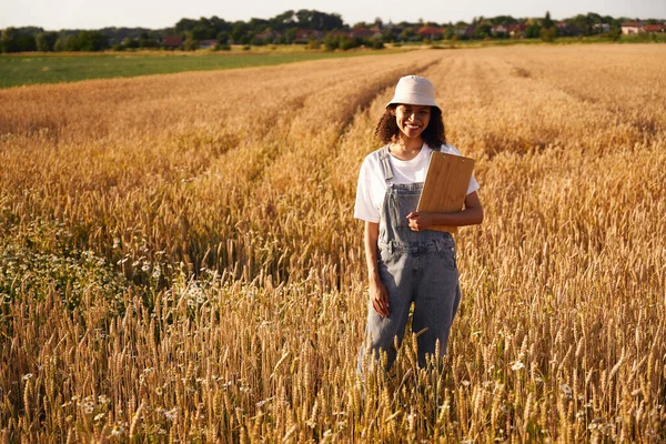 Woman agronomist standing with clipboard on wheat field background during harvesting