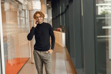 Gözlüklü gülümseyen erkek satış müdürü modern çalışma alanında telefonla konuşuyor.