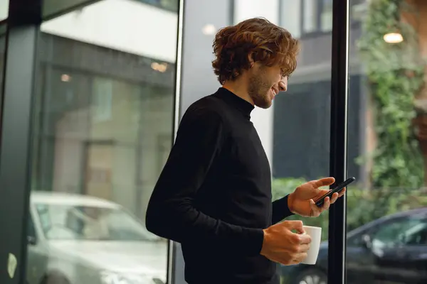 笑顔のビジネスマンは窓の近くに立っている間携帯電話を使用し コーヒーを飲む — ストック写真