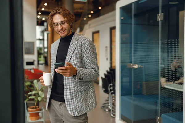 笑顔のビジネスマンは コワーキングの背景に立って携帯電話とコーヒーを飲む — ストック写真
