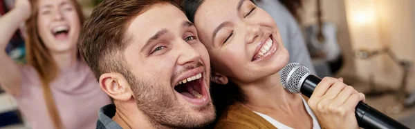 Bring the song to life. Young excited couple or friends holding microphone and singing together while playing karaoke with friends at home. Group of people having fun at karaoke home party