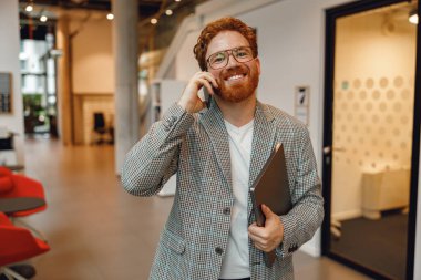 Bilgisayarlı gülümseyen girişimci pencerenin yanındaki modern ofiste telefonla konuşuyor