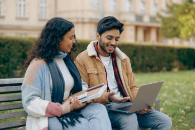 Mutlu öğrenciler bankta oturup dizüstü bilgisayara bakıyorlar ve projelerini tartışıyorlar.
