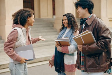 Genç öğrenciler kampüsün yanında durup kitap tutarak konuşuyor ve gülüyorlar. 
