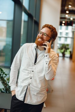 Gözlüklü, gülümseyen erkek yönetici modern çalışma alanında telefonla konuşuyor.