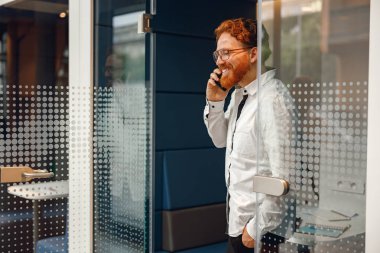 Gözlüklü, gülümseyen erkek yönetici modern çalışma alanında telefonla konuşuyor.
