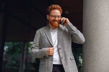 Gözlüklü, sakallı, serbest çalışan bir erkek iş yerinde telefonla konuşuyor.