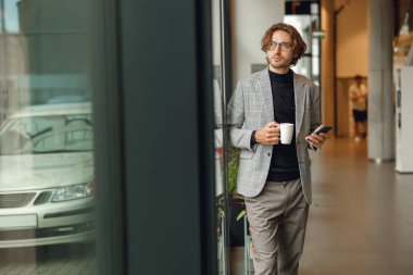 Gülümseyen işadamı pencerenin yanında dururken cep telefonu kullanır ve kahve içer.