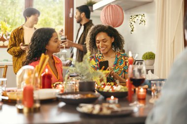 Gülümseyen genç arkadaşlar evde tatil partisinde şarap içip telefonlarına bakarlar.