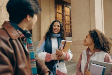 Üniversite kampüsünün yanında elinde kitaplarla dizüstü bilgisayarla birbiriyle konuşan mutlu öğrenciler.