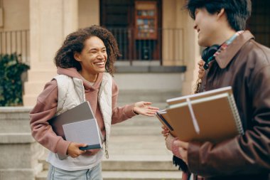 Üniversite kampüsünün yanında elinde kitaplarla dizüstü bilgisayarla birbiriyle konuşan mutlu öğrenciler.