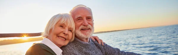 Romantic boat tour. Portrait of beautiful smiling senior couple holding hands, hugging and relaxing together while sitting on the side of sailboat or yacht floating in sea on a sunny day