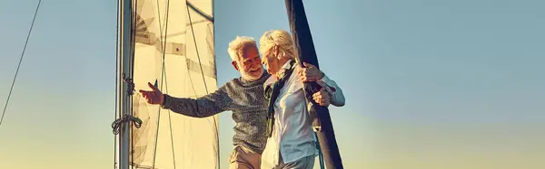 Happy senior man pointing at the horizon while standing with his wife on the side of a sail boat or yacht deck floating in a calm blue sea, enjoying sunset and amazing view, travelling together