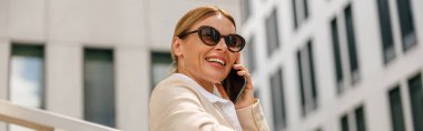 Smiling woman office worker is talking by phone while standing on modern building background