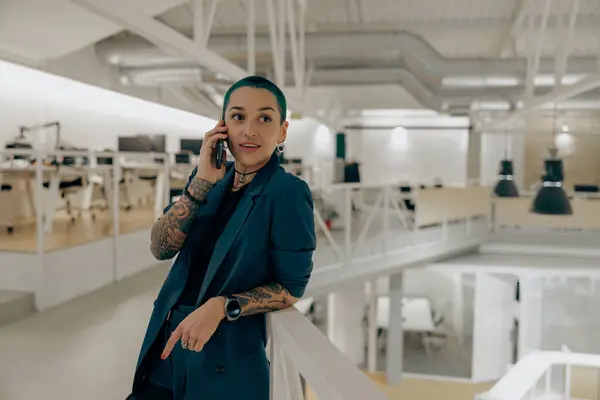Pretty tattooed female business owner is talking mobile phone while standing in modern coworking