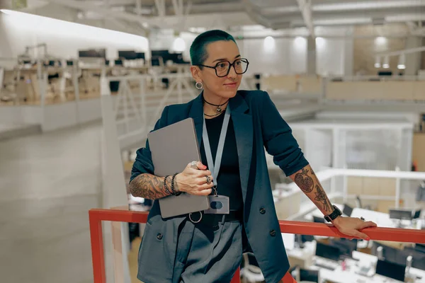 Creative tattooed business woman in glasses holding laptop and looking at side in modern office