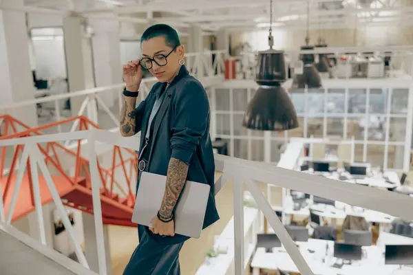 Focused tattooed business woman with green short hair holding laptop standing in modern office